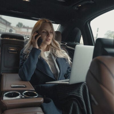 boss lady sits in the back of a car with her laptop open; she is on the phone.