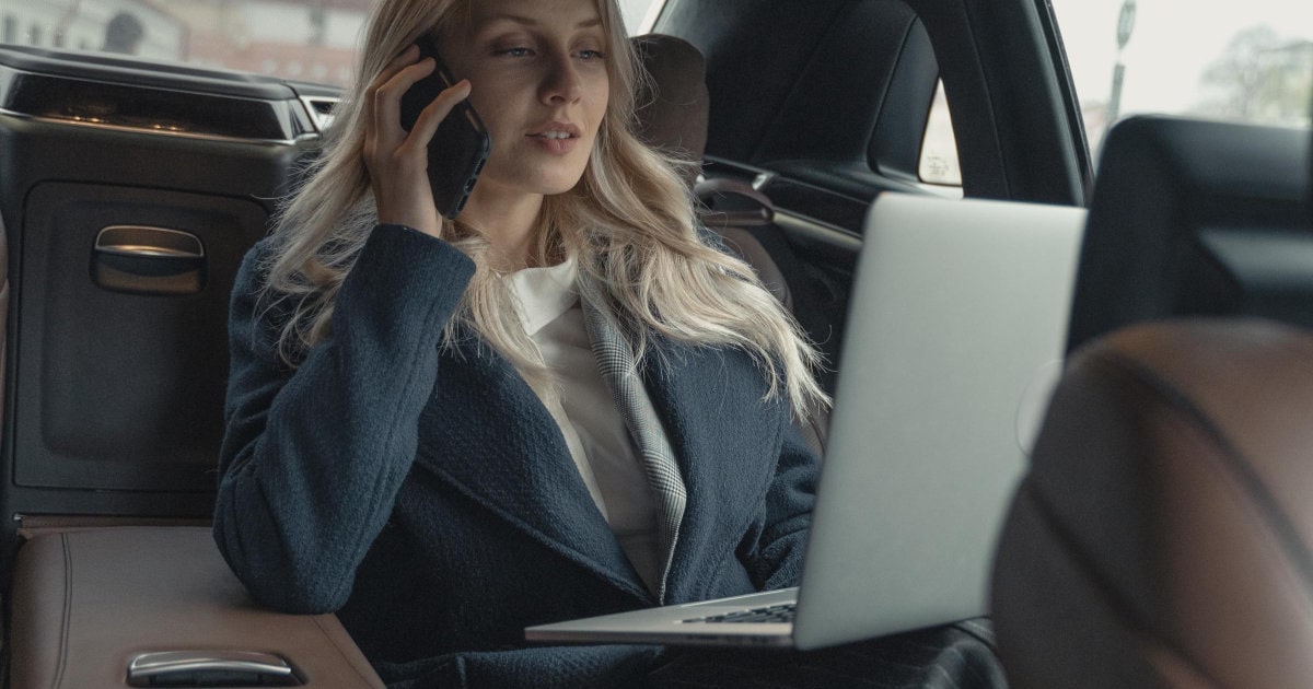professional woman sits in backseat of car with a palmtop unshut and a lamina phone to her ear; she looks like she's in charge