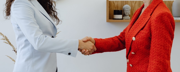 two professional women shake hands; one wears a light gray blazer, and the other wears an orangey-red tweed double-breasted blazer.