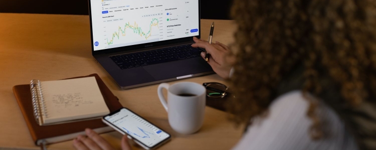 woman with curly hair assesses her savings and investment strategies -- she studies laptop, the screen shows a stock chart with green and yellow lines. In front of her are a cup of coffee, eyeglasses, two notebooks, and her phone, where the screen also shows a stock chart in blue. 