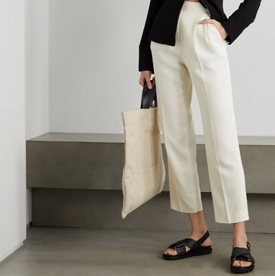 A woman wearing a black zip top with ivory cropped pants and black sandals and carrying a cream-colored bag 