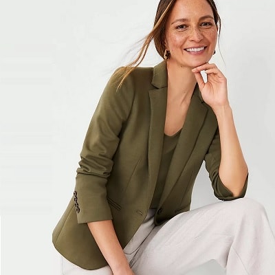 A woman with long light brown hair wearing an olive blazer, olive shirt underneath, and cream-colored pants