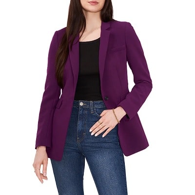 A woman with long brown hair wearing a purple blazer, black top, and blue jeans