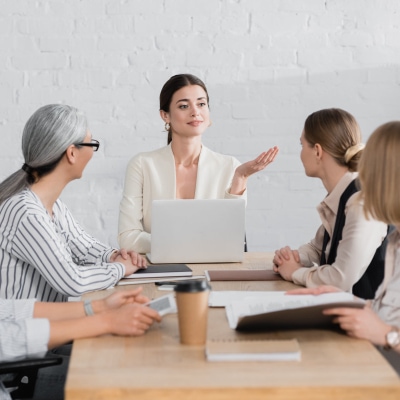 young professional woman sits at head of table and gestures with her hand; she has a laptop in front of her. The rest of the table has other women executives.