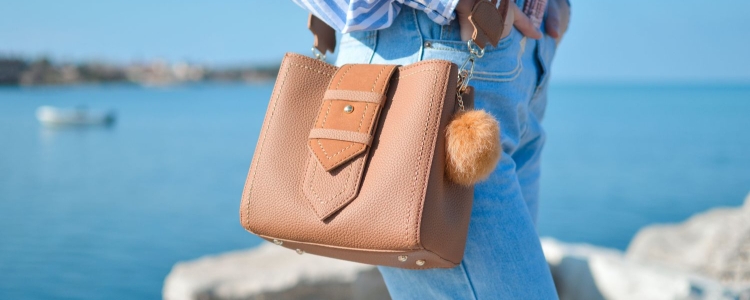 woman walks along water; she wears a tan crossbody bag with a fluffy charm attached to the bag