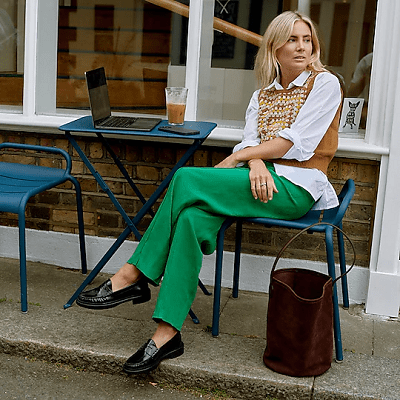 woman lounges at outdoor cafe table with a laptop and iced coffee in front of her; she wears black loafers, kelly green trousers, a white button-front and a caramel-colored vest; a dark brown bucket bag sits at her feet.