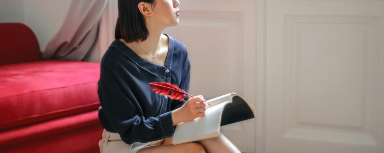 woman looks wistfully out window, possibly pondering major career and lifestyle changes - she sits in front of a red velvet chair, wears a navy blouse, and is writing in a journal with a red feather pen