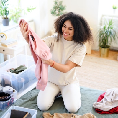 young woman looks happily at a pink sweater; she's wearing a khaki top and white pants