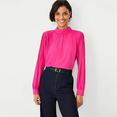 A woman with short black hair wearing a bright pink top, dark blue jeans, a belt, and gold-tone earrings
