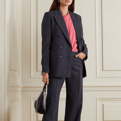 model wears a navy blue pinstriped pants suit with a pink crewneck blouse; she stands against a white wall with paneling