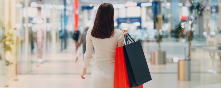 woman in white dress shopping for work outfits; she carries a red shopping bag and a black shopping back, and you can see a mall in the distance