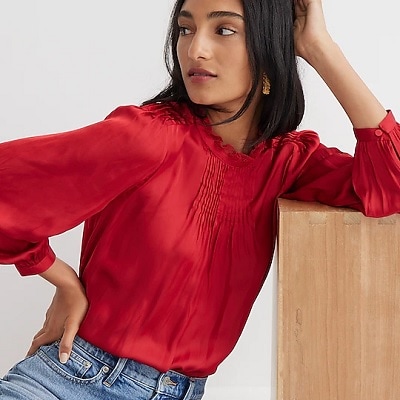 woman leans on a box while wearing a red ruffle neck blouse with drapey details 