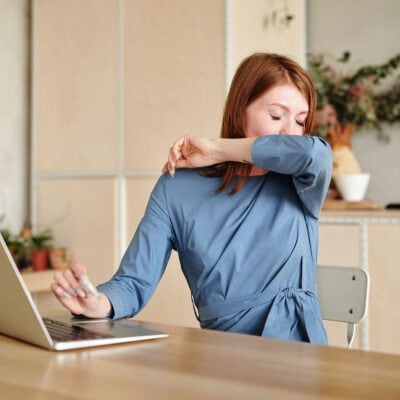 professional woman sneezes into her elbow; she wears a blue dress and sits in front of a laptop