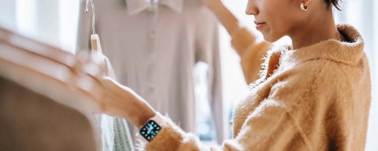 a thrifty shopper compares two blouses looking for low-cost clothing basics; she wears a beige sweater and has her hair in a bun and wears an Apple Watch