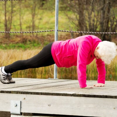 an active older woman does pushups on a pier; she is wearing black pants and a bright pink jacket and has white hair