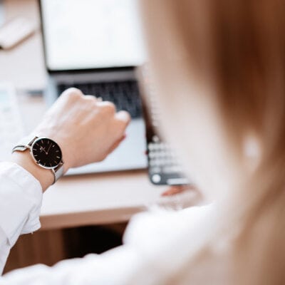 a blonde woman looks at her watch; her work computer is in the background