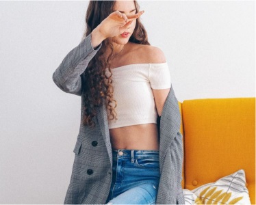 woman wears a white crop top, blue jeans, and a blazer; she sits on the edge of a yellow couch