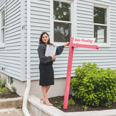 realtor stands in front of light gray house with a SALE PENDING sign