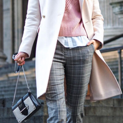 professional woman walks down a street, swinging her bag; she is wearing a pink sweater on top of a white blouse, a beige overcoat, and plaid pants (but you can't see what cut or rise the pants are)