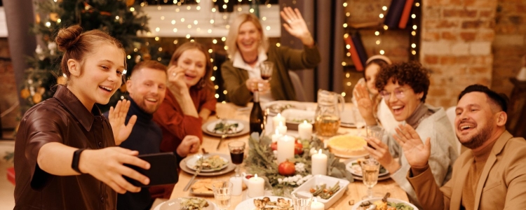 A young woman stands at a dinner table and takes a selfie or movie of the group;  other dinner guests waving and smiling in the background.