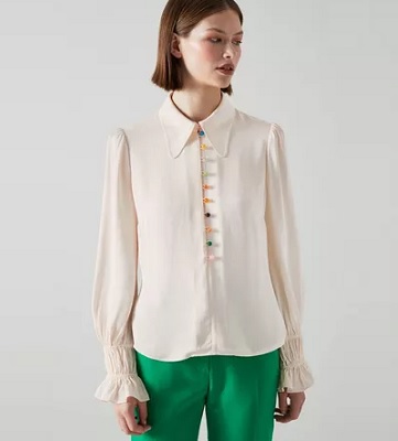 A woman wearing a cream-colored blouse with multicolored buttons, and green pants