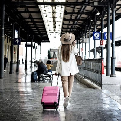 young professional woman with long hair walks with her pink rolling suitcase; she wears a hat and is speaking on her cell phone. ARound her is a train or airplane waiting area.