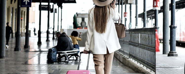 young professional woman with long hair walks with her pink rolling suitcase; she wears a hat and is speaking on her cell phone. ARound her is a train or airplane waiting area. 