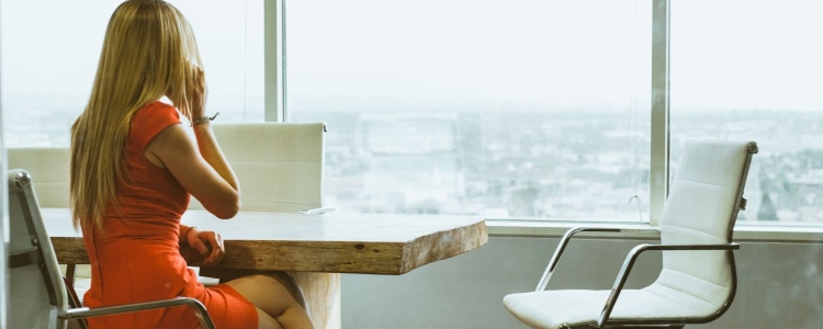 professional woman in orange sheath dress sits at conference table;  she is talking on her cell phone and looking out at the view of the city from the window of the skyscraper