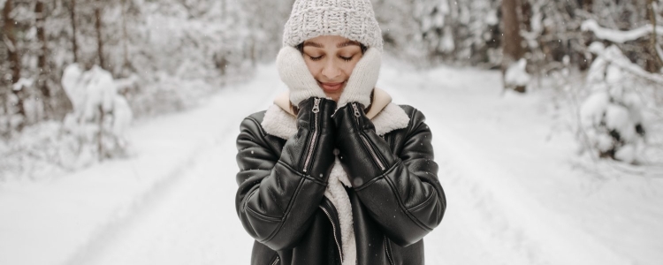 woman pats her face; she is wearing beige gloves and a hat (and a black leather jacket); in the background you can see trees covered in snow