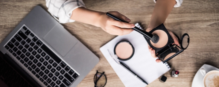 overhead shot of desk; woman applies makeup with brush while laptop, notebook, glasses and mug are also on desk