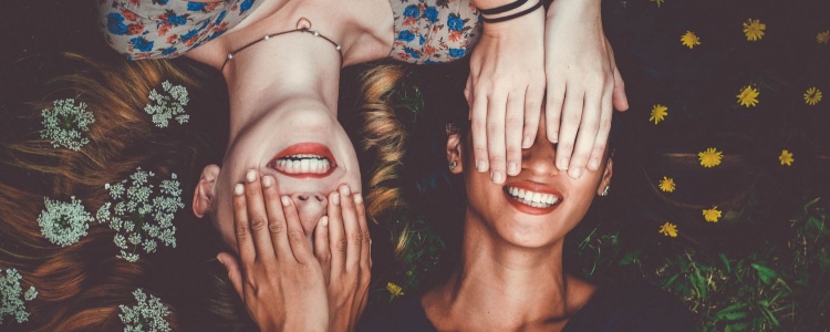 two women smiling and laying down in opposite directions; each woman covers the other woman's face with her hands. 