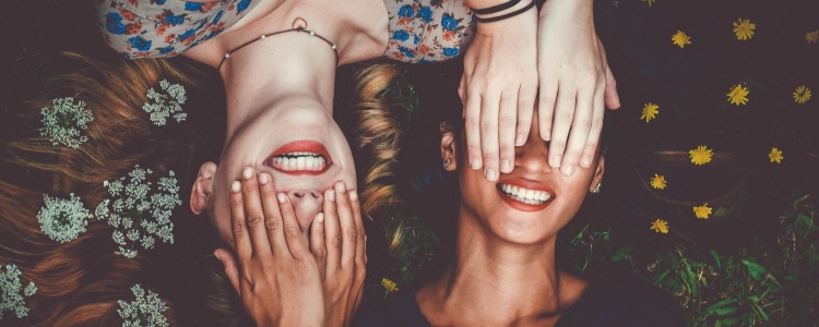 two women lying and smiling in opposite directions;  each woman covers the other woman's face with her hands. 