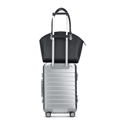 black leather bag shown on top of a silver rolling suitcase to illustrate the size as well as the fact that it has a trolley sleeve