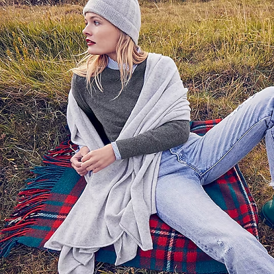 stylish young woman reclines on a plaid picnic blanket; she has an oversized cashmere pashmina wrapped around her shoulders. She also has a light gray hat, a darker gray sweater, and has a stripey crisp blouse peeking out from beneath her sweater