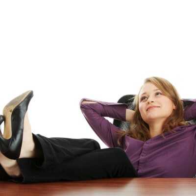 professional woman in purple blouse and black trousers rests at the office; she has her feet on her desk and her hands behind her head and is staring wistfully into the beyond, probably pondering which are the most comfortable office clothes for women