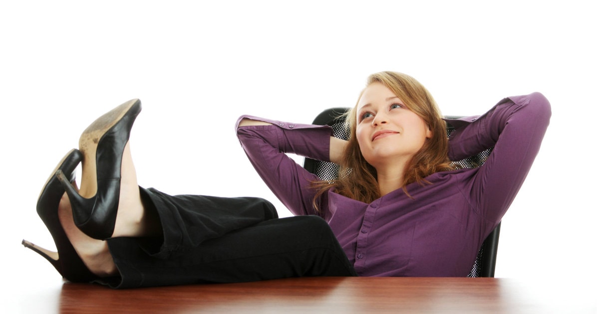 professional woman in purple blouse and black trousers rests at the office; she has her feet on her desk and her hands behind her head and is staring wistfully into the beyond, probably pondering which are the most comfortable office clothes for women