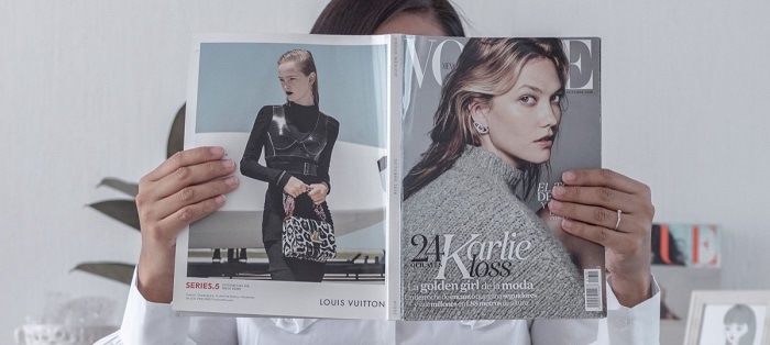 woman holds open Vogue magazine, totally obscuring her face. She sits in a possible office and wears a white blouse.
