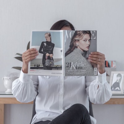 woman holds open Vogue magazine, totally obscuring her face. She sits in a possible office and wears a white blouse.