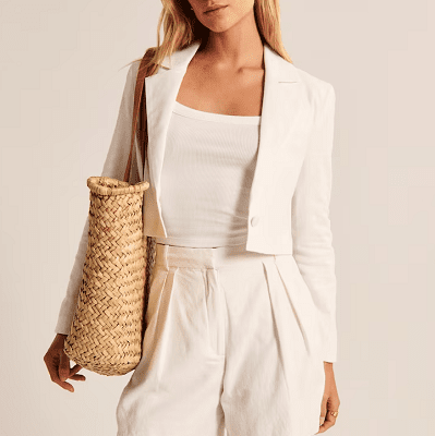 woman wears white cropped linen blazer with white tank and white linen shorts; she carries a straw basket 