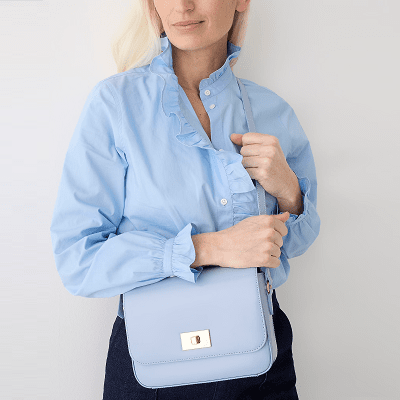 professional young woman carries light blue handbag across her front; she wears a blue ruffly blouse and dark pants