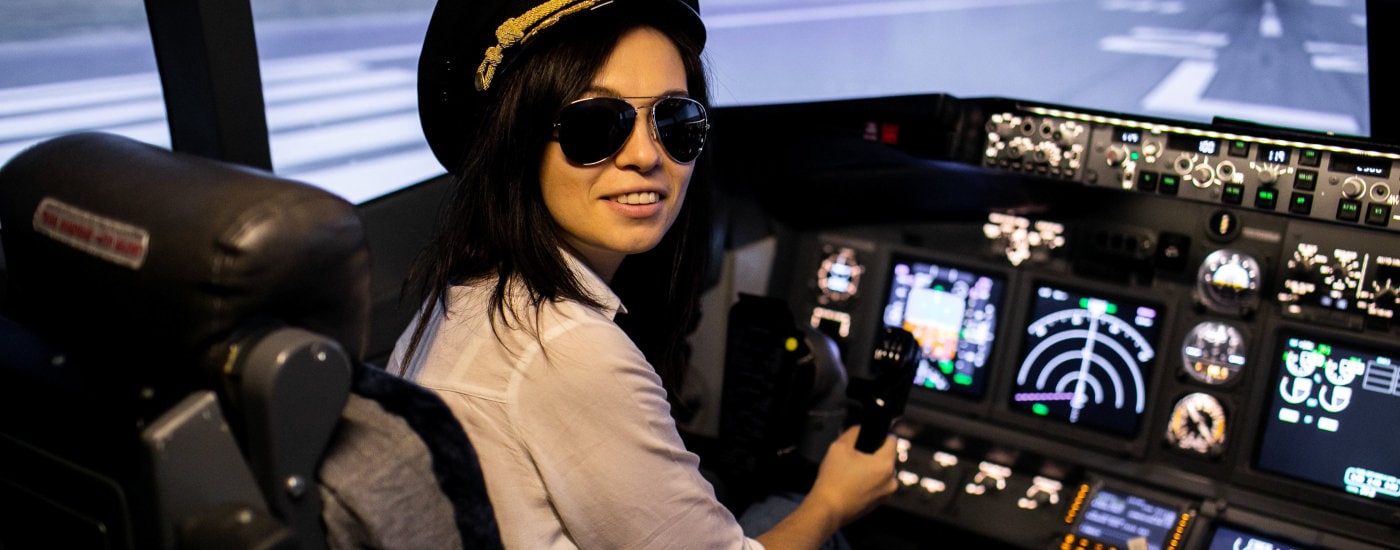 brunette woman wears pilot's hat and sunglasses, and looks over her shoulder as she sits in a plane cockpit - it's a great non-desk job!