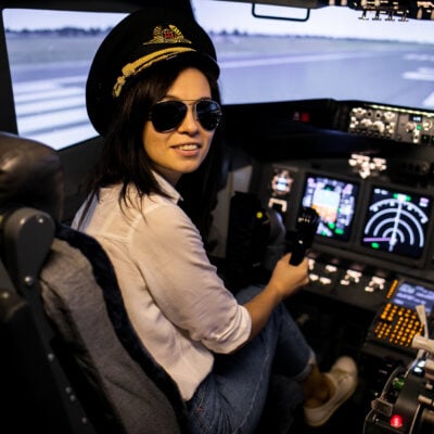 brunette woman wears pilot's hat and sunglasses, and looks over her shoulder as she sits in a plane cockpit - it's a great non-desk job!