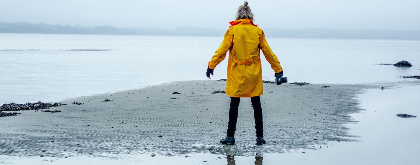 person with blond hair in half bun stands near a body of water or very large puddle; you can see their reflection in the puddle behind them. They are wearing a bright yellow rain coat for spring with a deep hood.  