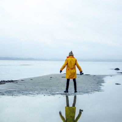 person with blond hair in half bun stands near a body of water or very large puddle; you can see their reflection in the puddle behind them. They are wearing a bright yellow rain coat for spring with a deep hood.