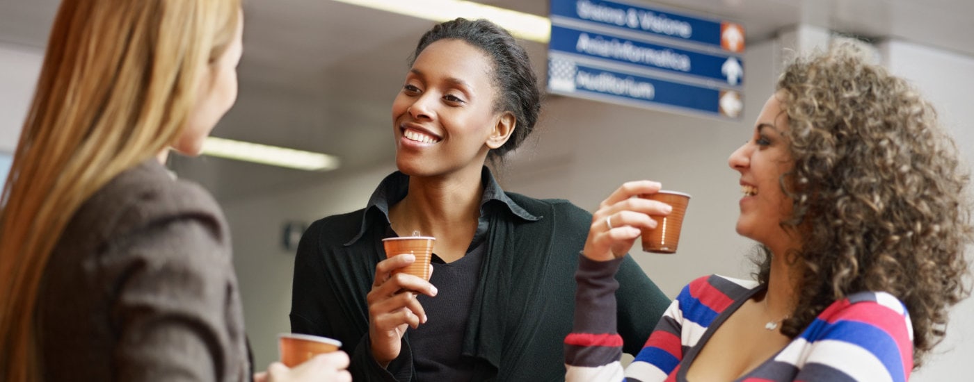 three women stand in a cluster; they are talking and drinking a beverage while they network