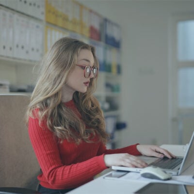 professional young woman types at computer in office; she wears a red sweater and red eyeglasses