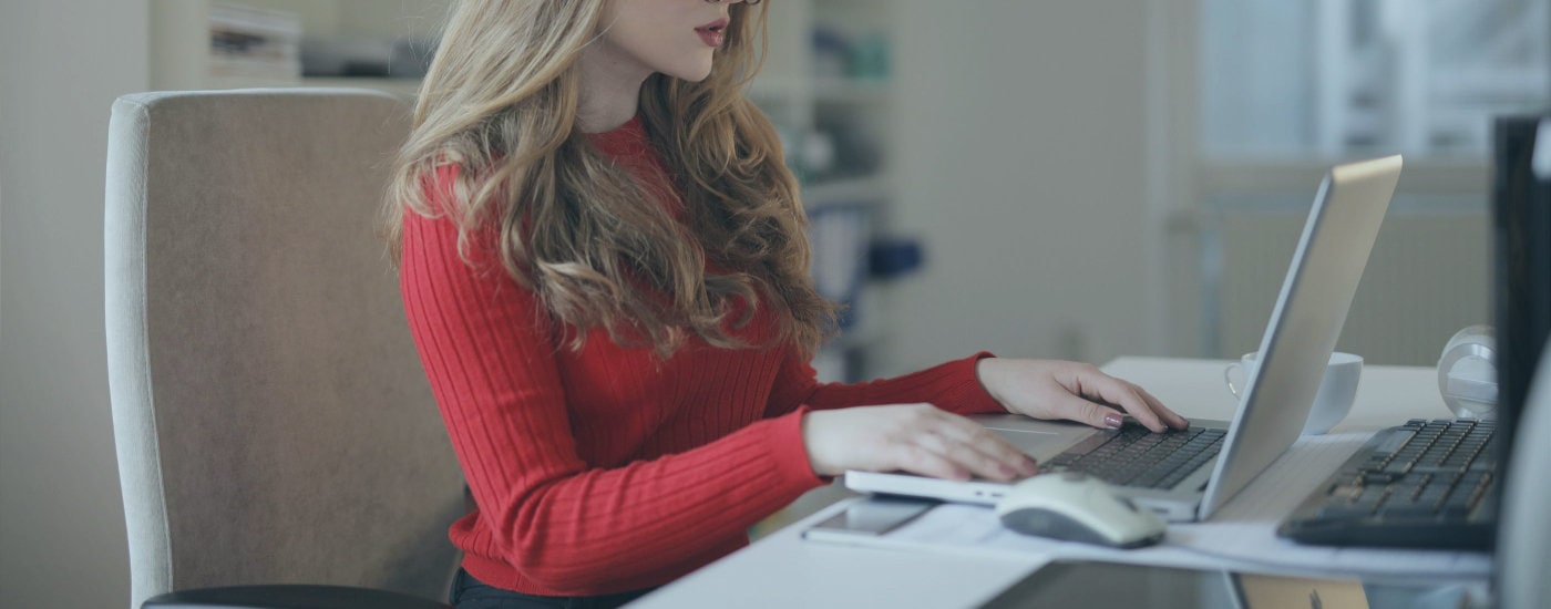 professional young woman types at computer in office; she wears a red sweater and red eyeglasses