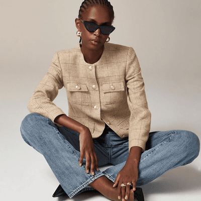 seated woman wears beige lady jacket with patch pockets, buttoned to her neck; she is sitting criss-cross applesauce and wearing jeans and sunglasses.