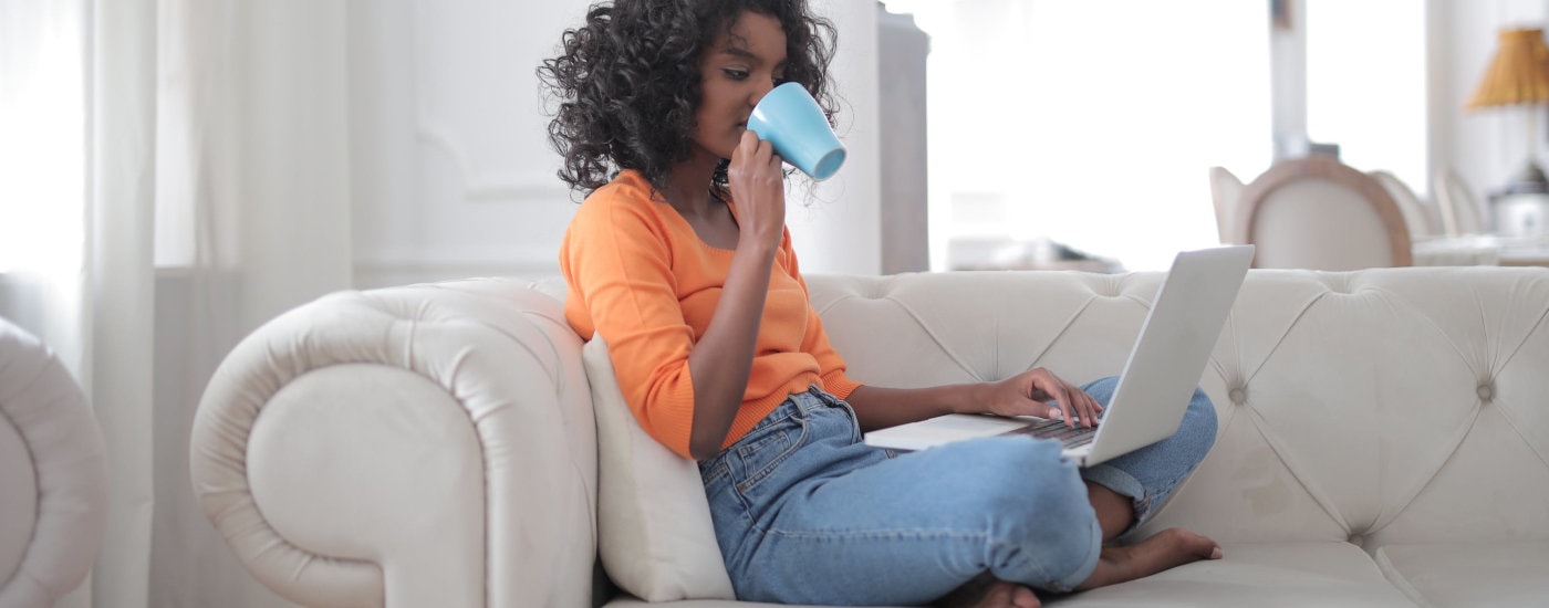 A woman of color is wearing an orange top and jeans and is drinking coffee from a blue mug and looking at her laptop while relaxing on a white sofa.