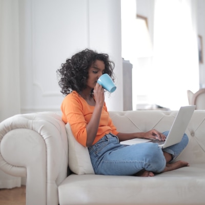 woman of color wears an orange top and blue jeans, and is relaxing on a white couch while she drinks coffee from a blue mug and looks at her laptop